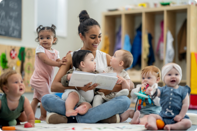 An educator smiles and sits on the floor of a classroom, holding two babies. One baby stands up and holds the educator's shoulder, and three others sit on the floor beside her. An educator smiles and sits on the floor of a classroom, holding two babies. One baby stands up and holds the educator's shoulder, and three others sit on the floor beside her.