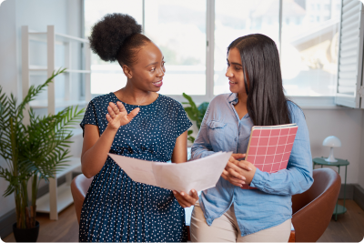 A coach with notes in their hand, talking to and standing next to an educator holding a folder. A coach with notes in their hand, talking to and standing next to an educator holding a folder.