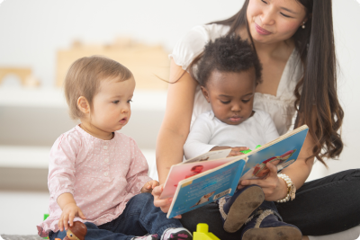A teacher is sitting on the floor reading to a small child in her lap and another small child sitting next to her right. A teacher is sitting on the floor reading to a small child in her lap and another small child sitting next to her right.