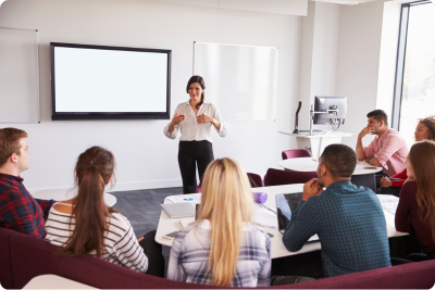 Trainer facilitating a course, in front of a whiteboard, for several educators in a small lecture room.