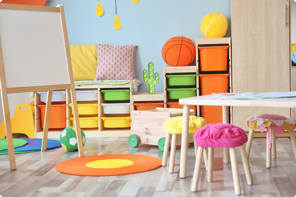 A classroom, featuring a whiteboard, a table with stools, a rug, a toy cart, balls, and pillows on top of cubbies. A classroom, featuring a whiteboard, a table with stools, a rug, a toy cart, balls, and pillows on top of cubbies.